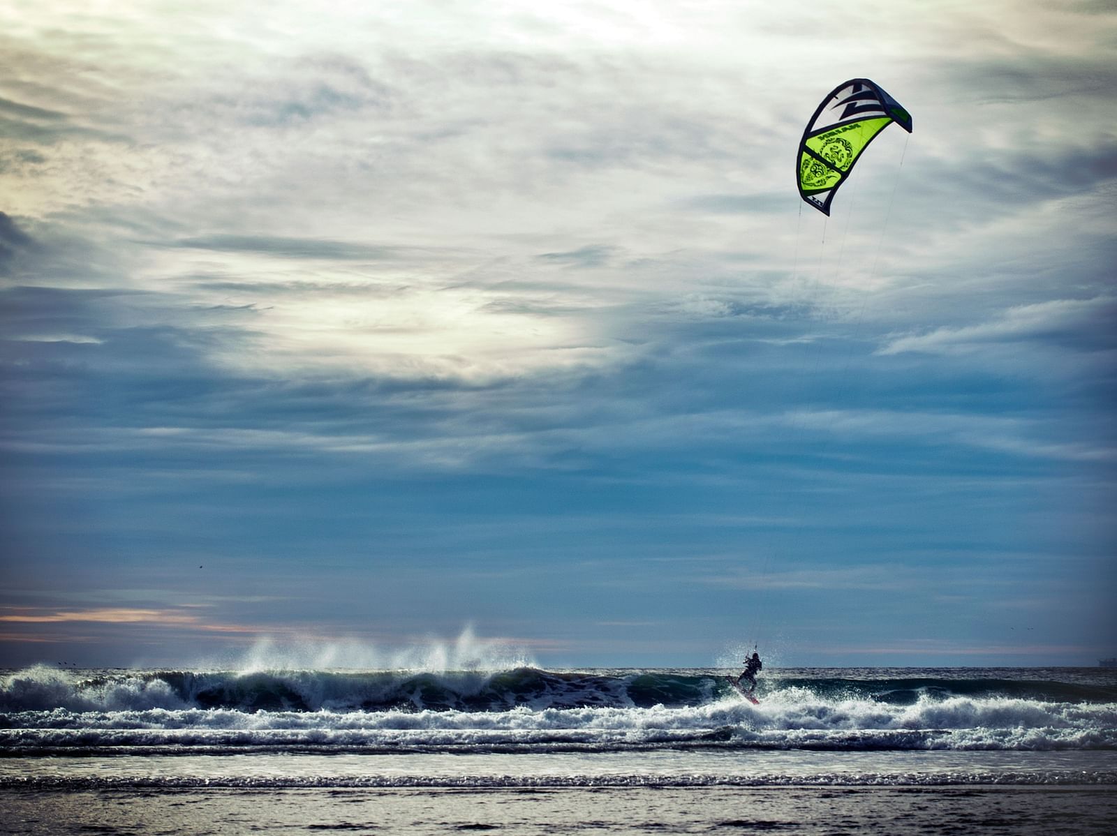 A brilliant shot of a person kite surfing on the beach with waves and white clouds in view.