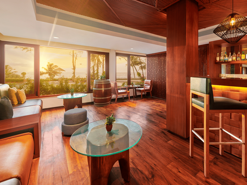 A coffee table in the centre with a leather sofa on the side, and a floor to ceiling window having sunset views inside the Upper deck Bar at The Resort, Mumbai, a Couples Resorts in Mumbai.