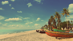 A sandy beach with boats and trees on one side and cloudy blue sky in the background.