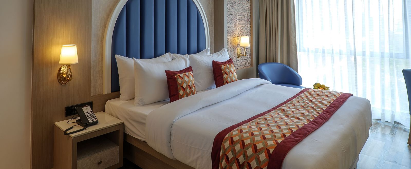 A well-lit hotel room features a bed with decorative pillows and a blue arched headboard, along with bedside tables - Melior Inn