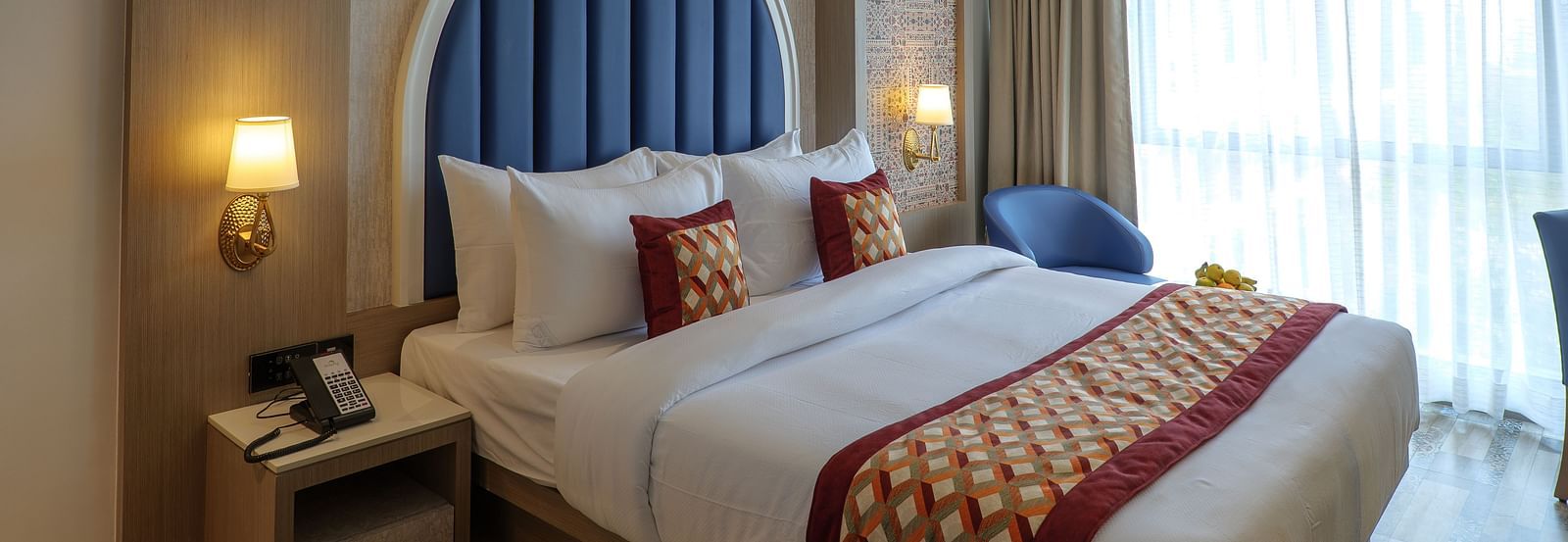 A well-lit hotel room features a bed with decorative pillows and a blue arched headboard, along with bedside tables - Melior Inn