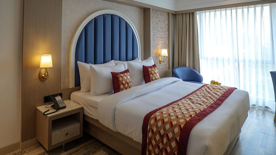 A well-lit hotel room features a bed with decorative pillows and a blue arched headboard, along with bedside tables - Melior Inn