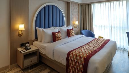 A well-lit hotel room features a bed with decorative pillows and a blue arched headboard, along with bedside tables - Melior Inn