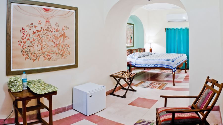 Hotel suite with a bedroom featuring a bed with blue and white striped bedding and teal curtains, a seating area with a wooden chair, a small table, a luggage rack, a mini-fridge, and artwork at Tijara Fort-Palace, Alwar.