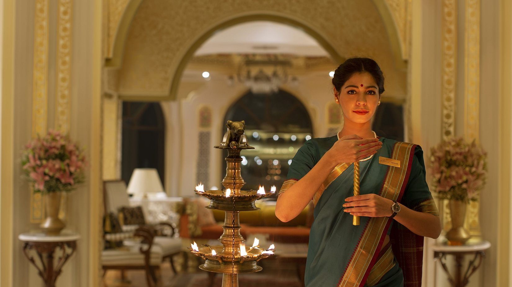 A woman in a saree lights a traditional lamp in a luxurious hotel lobby with golden accents - Heritage Village Resorts & Spa, Manesar