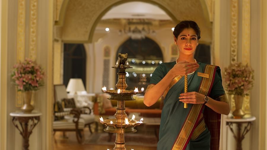 A woman in a green saree lights a traditional lamp in a luxurious hotel lobby with golden accents - Heritage Village Resorts & Spa, Manesar