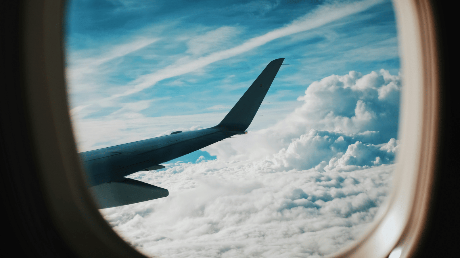 A view out of an aeroplane window showing part of the wing, with bright blue sky and fluffy white clouds below.