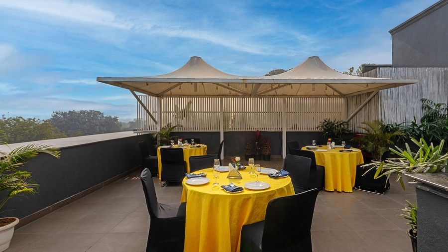 A rooftop dining area with two sheltered and others open air tables, featuring black chairs around yellow-clothed round tables, and plants lining the walls along the railing.