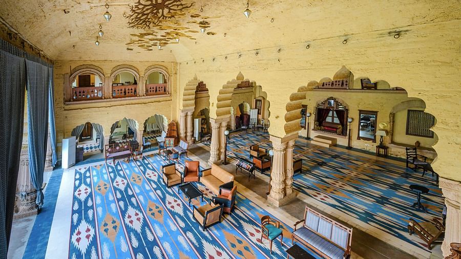 The interior of Tijara Fort-Palace - 19th Century, Alwar reveals a spacious hall with patterned carpets, arched doorways, seating areas, and decorative elements on the walls and ceiling.