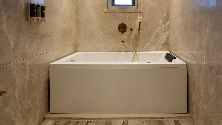A bright bathroom showcases a large white bathtub with tiled surroundings, a small window, and neutral-coloured wall tiles - Melior Inn