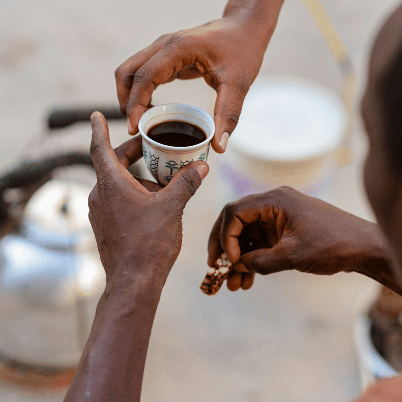 A close up of a small coffee cup being handed over to a person with a pot kept on the ground.