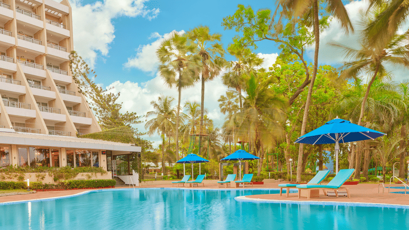 The poolside view of The Resort, Mumbai, featuring parasols, sun loungers, and towering trees.