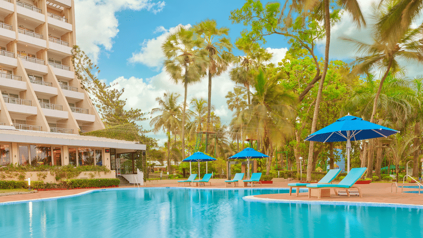 The poolside view of The Resort, Mumbai, featuring parasols, sun loungers, and towering trees.