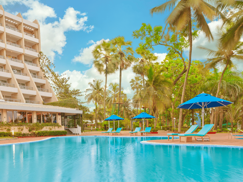 The poolside view of The Resort, Mumbai, featuring parasols, sun loungers, and towering trees.