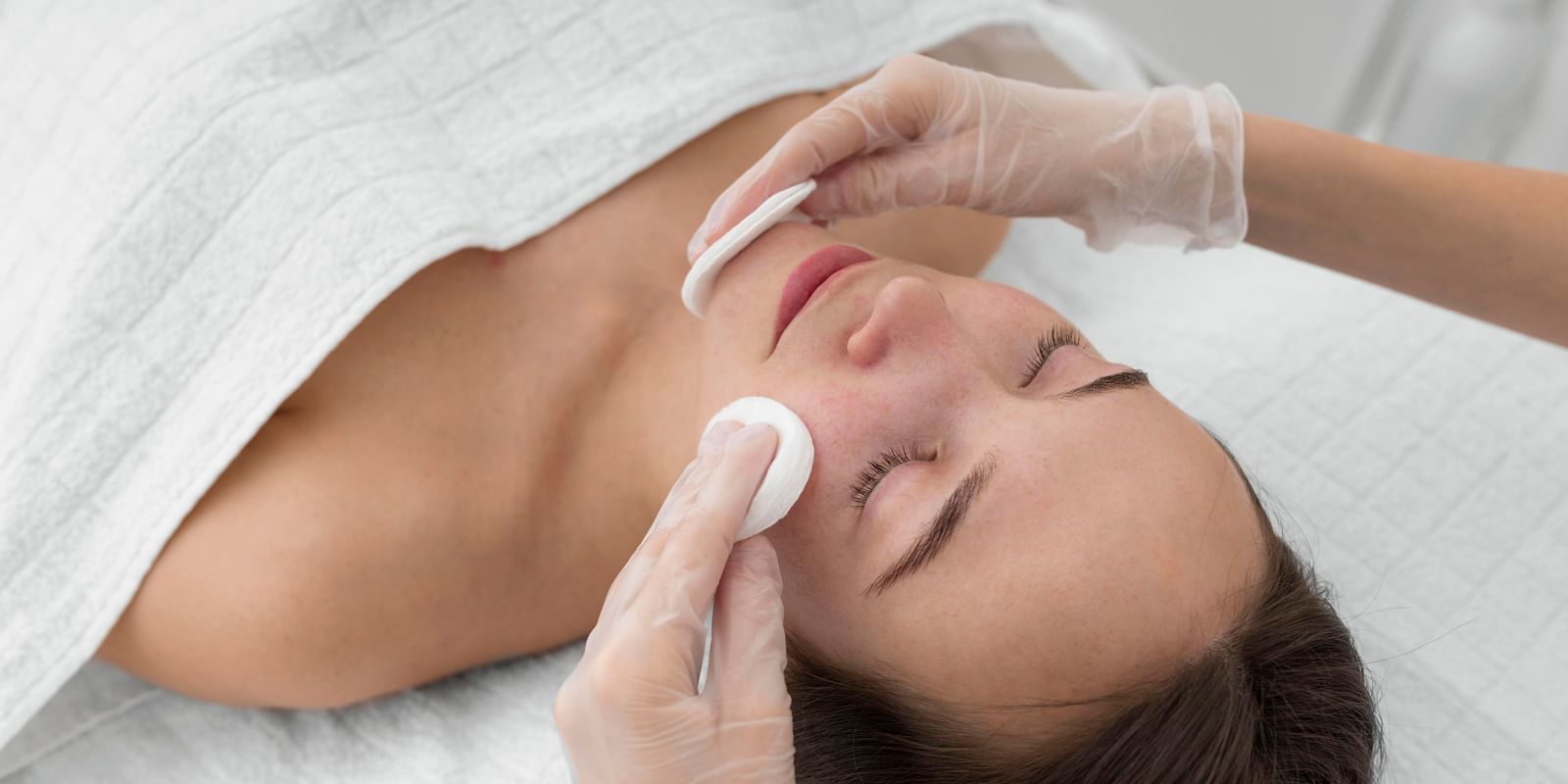 A beautician applying a cotton pad to a woman's face during a facial treatment.