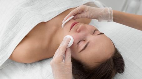 A beautician applying a cotton pad to a woman's face during a facial treatment.