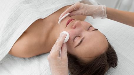 A beautician applying a cotton pad to a woman's face during a facial treatment.