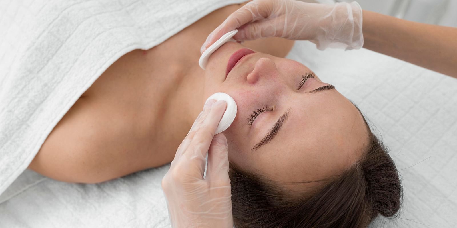 A beautician applying a cotton pad to a woman's face during a facial treatment.