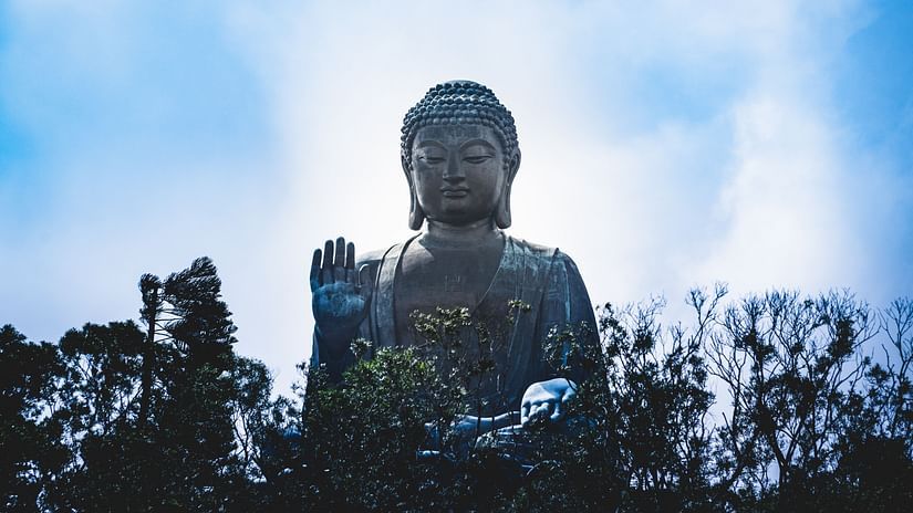 A massive statue of buddha with trees covering it and white clouds and blue sky in the background