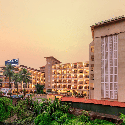 towering hotel buildings featuring balconies, a signage with hotel name, and surrounded by lush greenery, set against a golden hue filed sky - Golden Tulip, Candolim, Goa