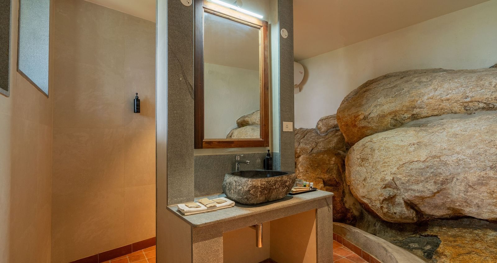 A stylish bathroom featuring a stone wall, modern fixtures, a large mirror, and a unique rock formation integrated into the design. | Mango Hill, Yercaud