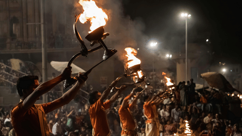 A nighttime shot of a religious ceremony (Ganga Aarti) with people holding flaming lamps and wearing traditional Indian clothing, with a large crowd in the background.