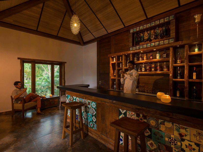 An overview of the seating arrangement with a bar counter and a staff member behind it and a customer sitting on the chair at Outpost the Bar at The Serai, Kabini.