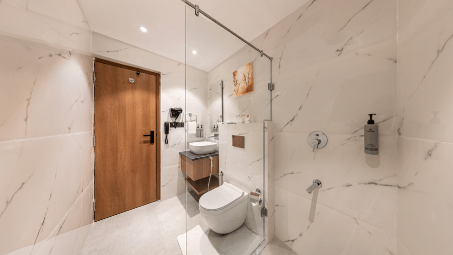 A modern bathroom interior at Lords Inn Katra featuring a white ceramic toilet, a clear glass shower partition, and polished marble walls.