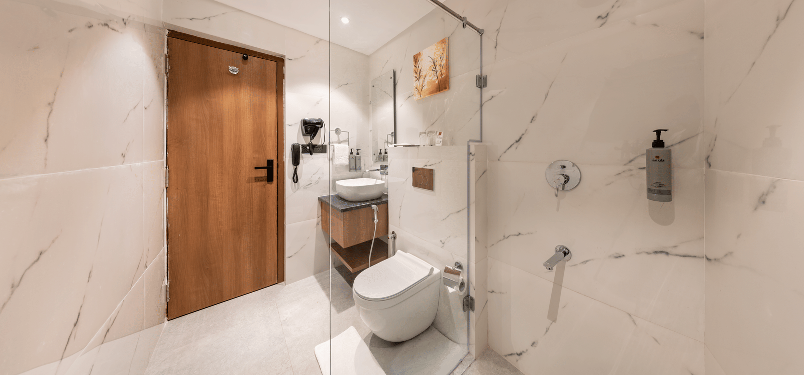A modern bathroom interior at Lords Inn Katra featuring a white ceramic toilet, a clear glass shower partition, and polished marble walls.