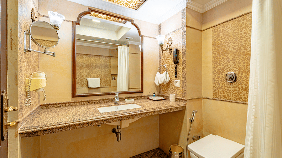 The bathroom of the Standard Room at Noor-Us-Sabah Palace, Bhopal, featuring a long beige marble vanity, a large arched mirror, and a clean, traditional layout.
