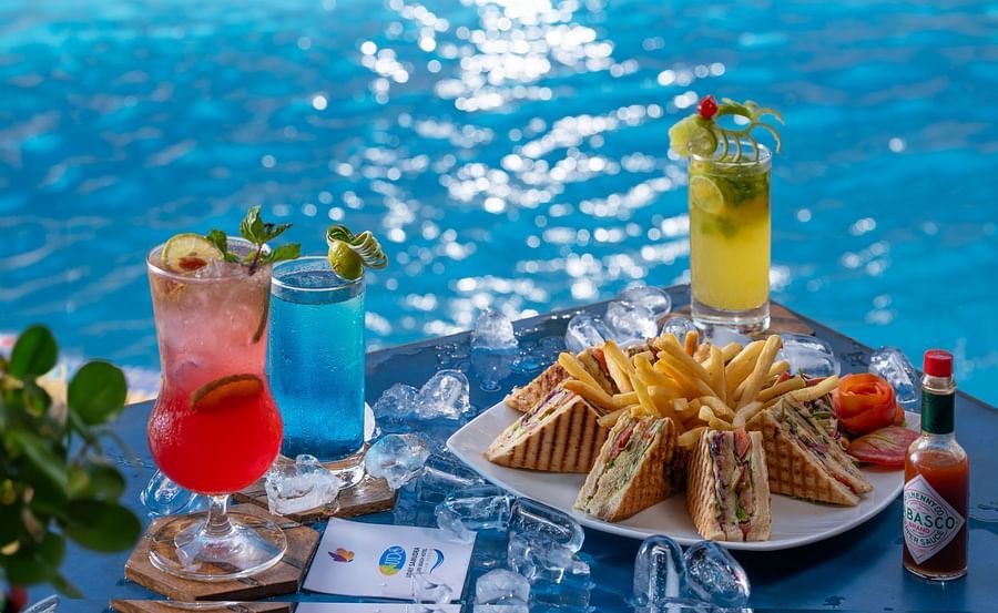 A table next to a sparkling blue pool holds a plate of club sandwiches and fries, surrounded by crushed ice and three colourful cocktails - Uday Samudra Leisure Beach Hotel.