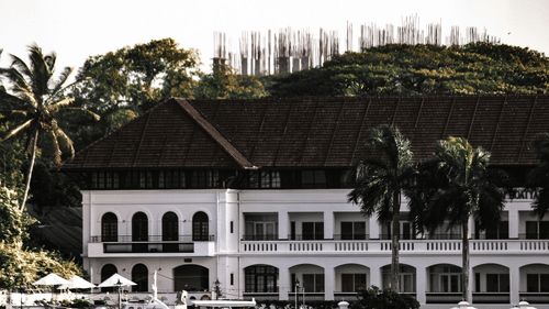 A white, colonial-style building with a dark roof and arches, next to water and palm trees in Kochi.