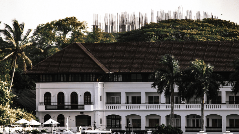 A white, colonial-style building with a dark roof and arches, next to water and palm trees in Kochi.