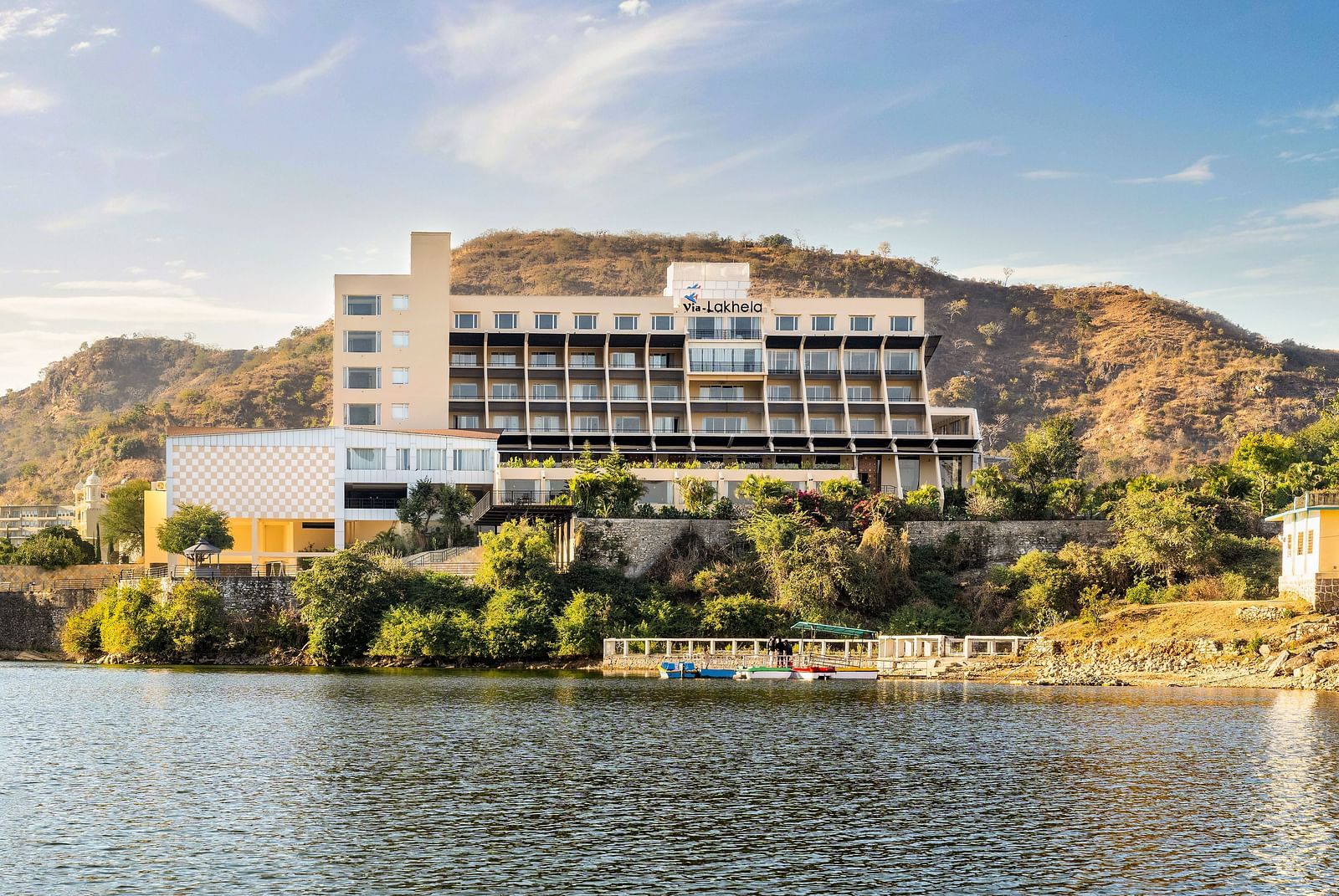 Hotel building by a lake with hills in the background under a bright sky - Via Lakhela Resort & Spa