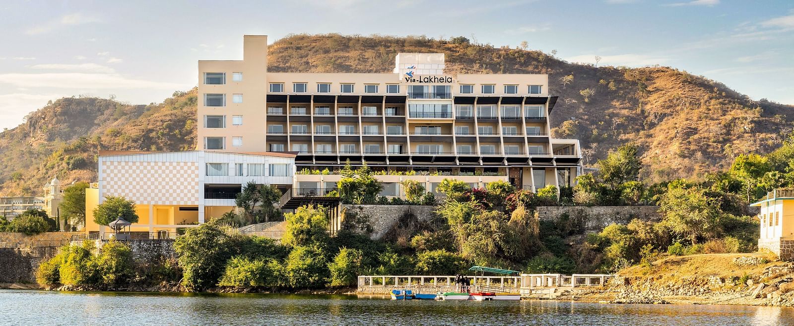 Hotel building by a lake with hills in the background under a bright sky - Via Lakhela Resort & Spa