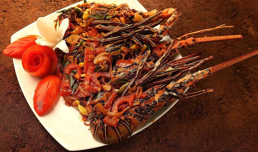 A cooked lobster dish, featuring visible claws and a seasoned body, is presented on a white plate with tomato and onion garnishes, set against a textured brown surface at Uday Samudra Leisure Beach Hotel.