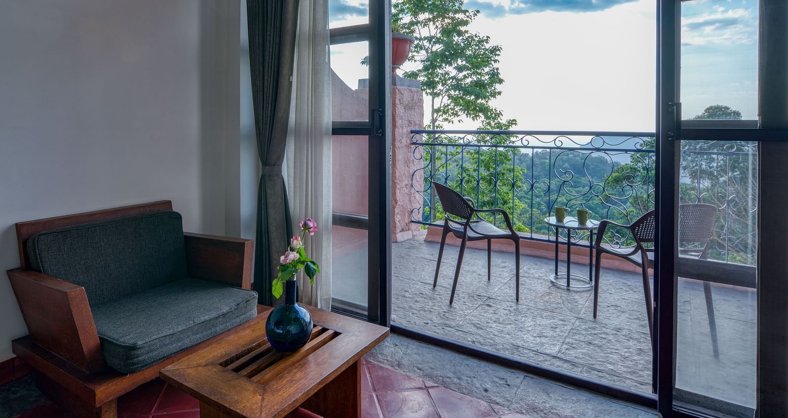 Seating area adjacent to glass doors opening to a balcony with outdoor furniture and a picturesque view of the surrounding landscape. | Mango Hill, Yercaud