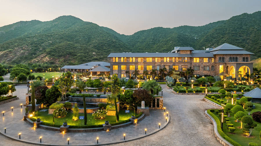 Resort exterior with illuminated building, circular driveway, landscaped gardens, pathways, and surrounding hills at dusk.