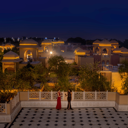 A couple stands on a spacious terrace overlooking a luxury resort illuminated by warm golden lights, surrounded by a serene ambience