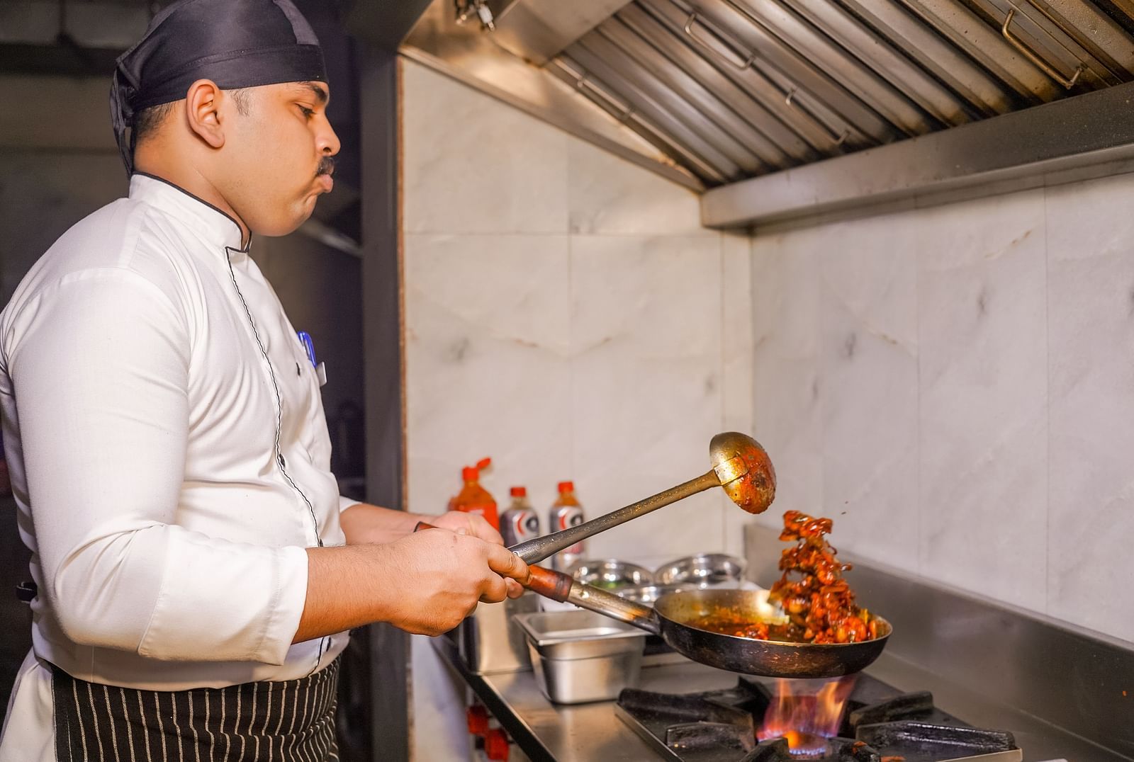 A chef cooking in a commercial kitchen with flames coming from the pan - TGI Ameya Grand, Goa