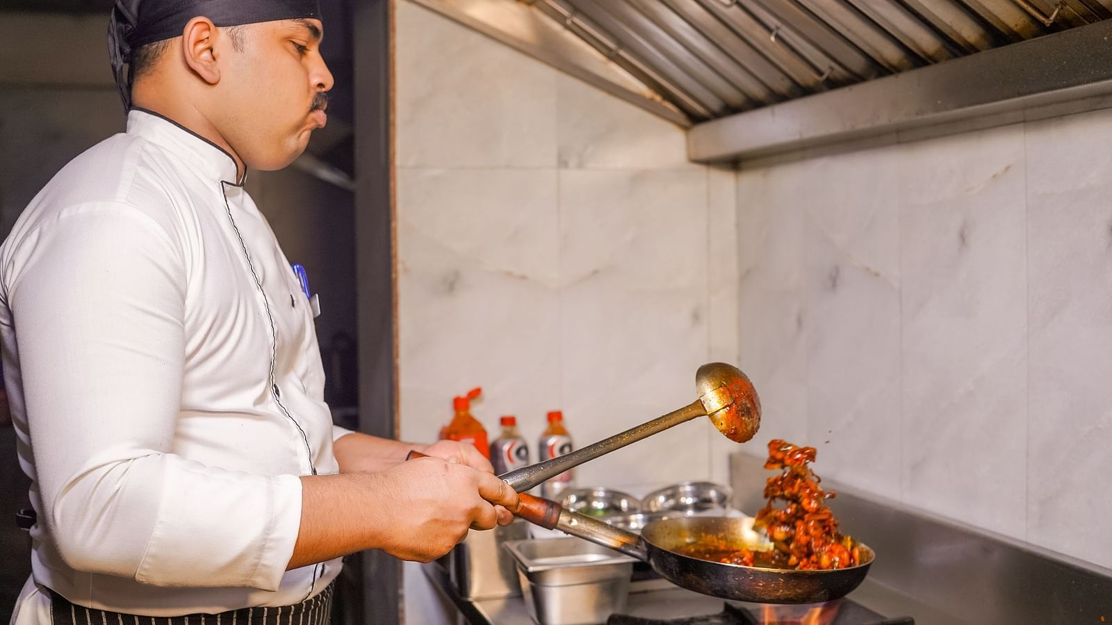 A chef cooking in a commercial kitchen with flames coming from the pan - TGI Ameya Grand, Goa