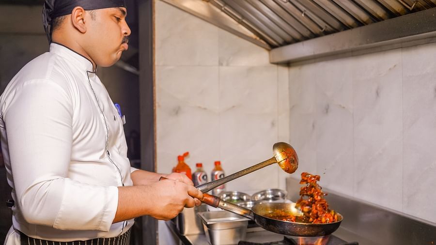 A chef cooking in a commercial kitchen with flames coming from the pan - TGI Ameya Grand, Goa