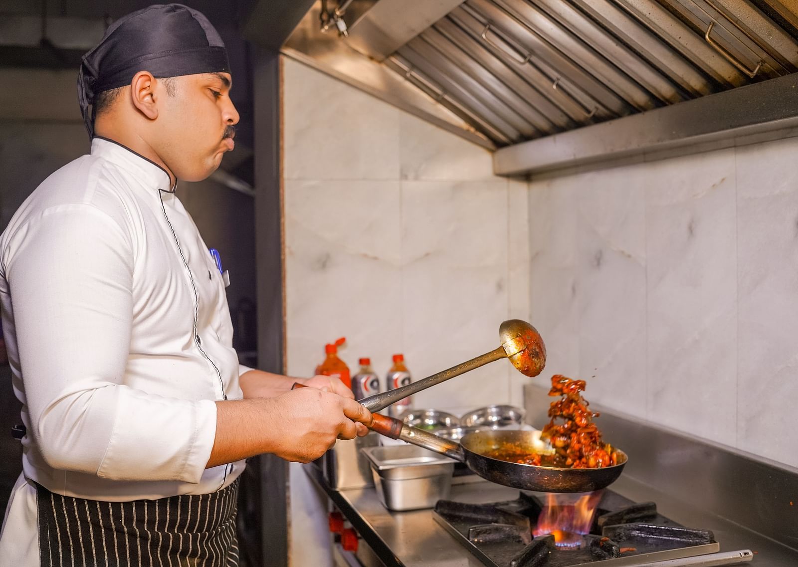 A chef cooking in a commercial kitchen with flames coming from the pan - TGI Ameya Grand, Goa