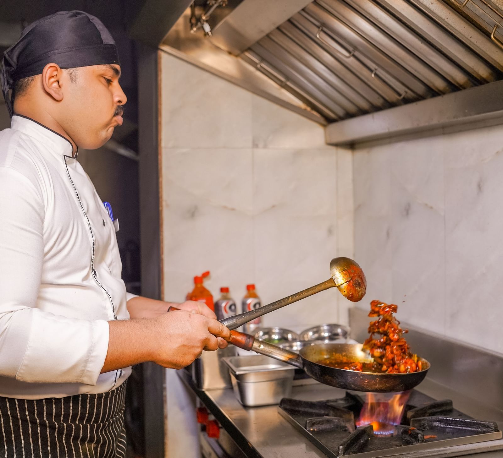 A chef cooking in a commercial kitchen with flames coming from the pan - TGI Ameya Grand, Goa