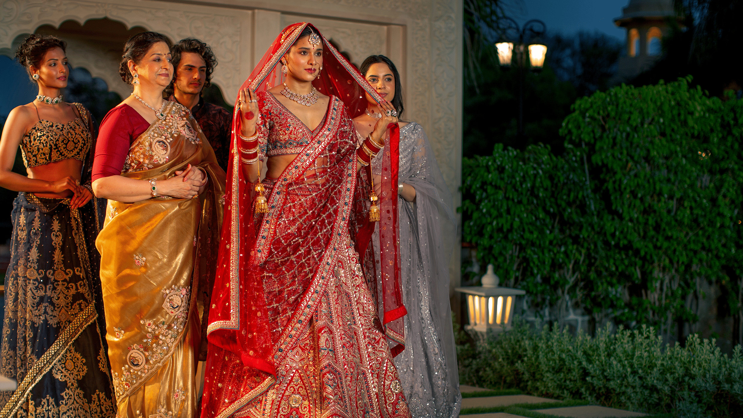 Four women in traditional indian sarees pose together at dusk, with soft lighting and a garden in the background.
