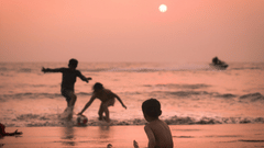 A silhouette of children playing in the shallow surf of a beach under a vibrant orange sky, with the sun low in the sky reflecting on the water.