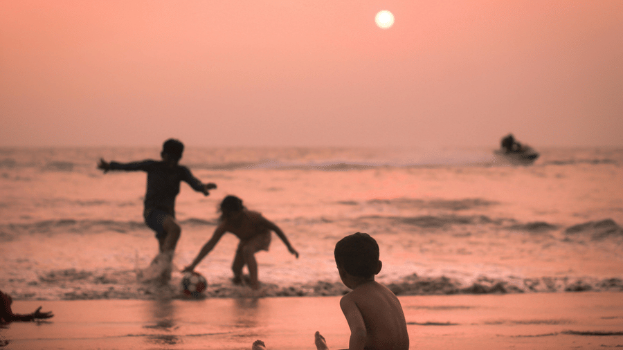 A silhouette of children playing in the shallow surf of a beach under a vibrant orange sky, with the sun low in the sky reflecting on the water.
