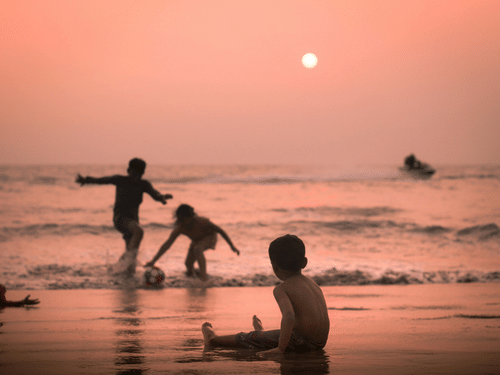 A silhouette of children playing in the shallow surf of a beach under a vibrant orange sky, with the sun low in the sky reflecting on the water.