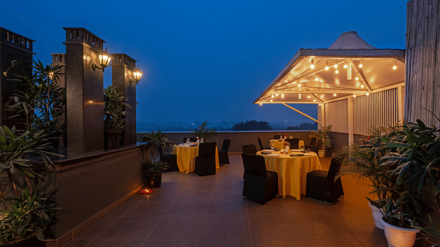 This rooftop dining spot offers sheltered access decorated with yellow string lights, with round tables draped in yellow and paired with black chairs, complemented by plants along the wall opposite the railing.
