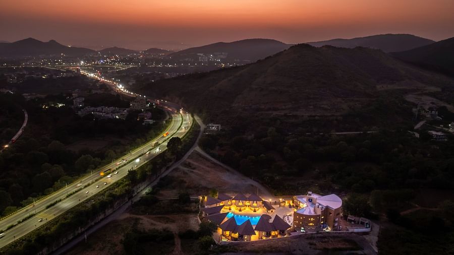 An aerial view of The Qualia Resort Club & Brewery, Udaipur at sunset, with mountains in the background and a road leading to the resort.